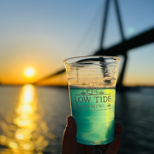 low tide brewing glass with teal drink held in front of sunset
